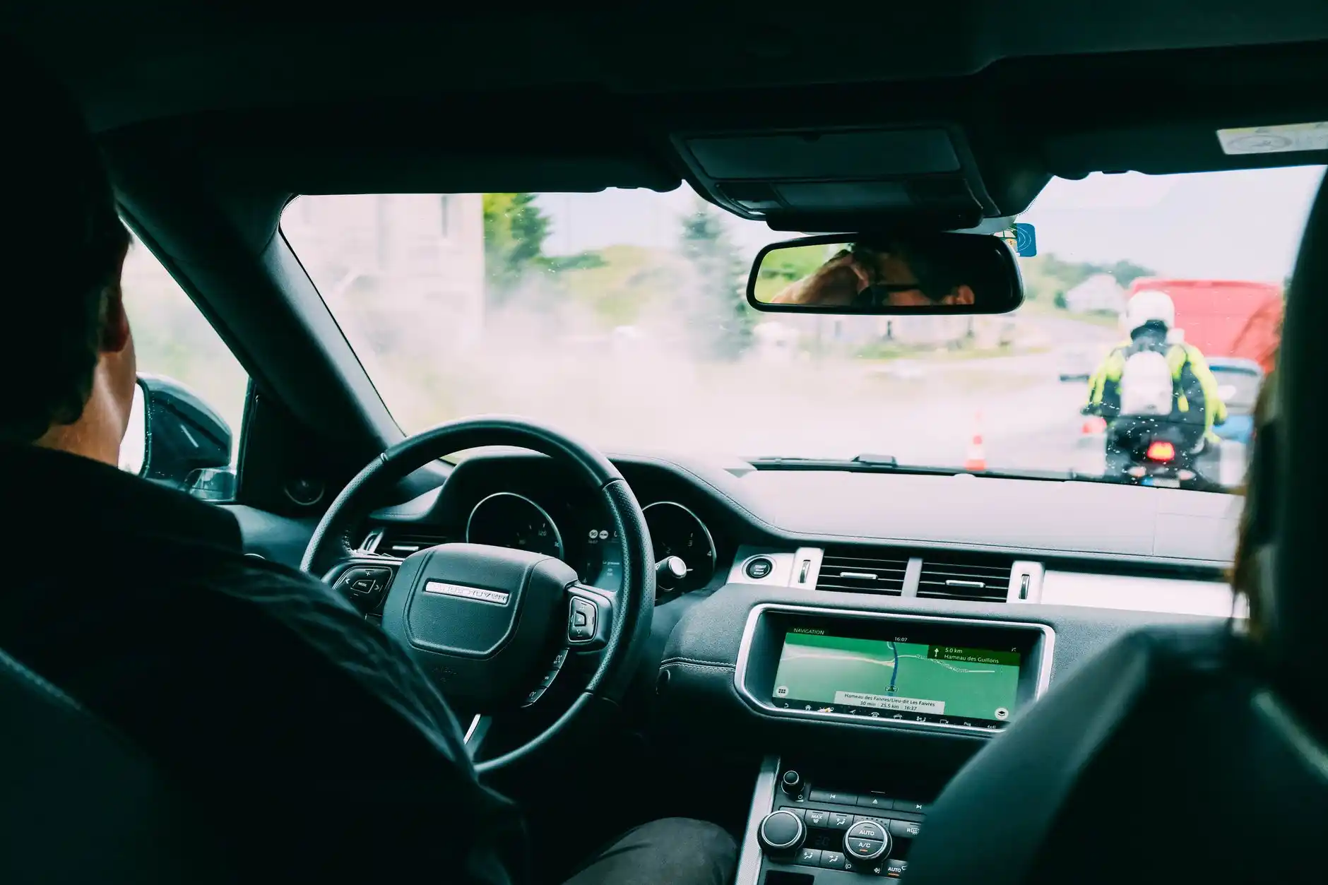 Interior view from a luxury SUV approaching SeaTac Airport, digital navigation showing arrivals access, surround-view camera active, rain droplets on windshield and evening traf...